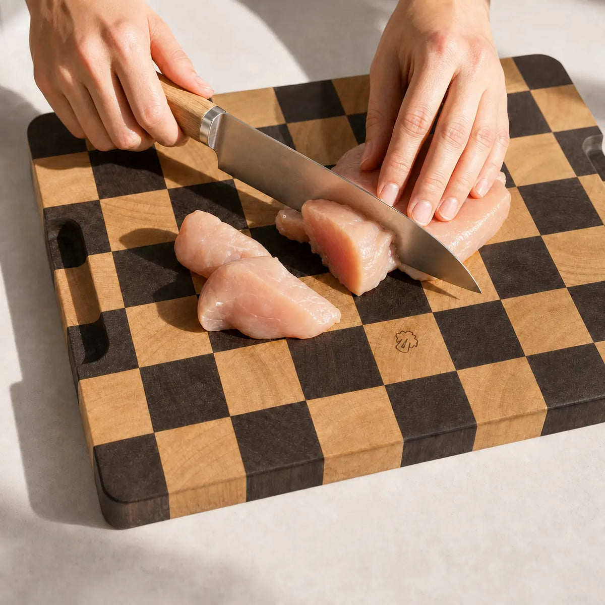 Close-up of Alorivo checkerboard end-grain wooden cutting board being used to slice raw chicken breast with a chef knife under bright, warm natural sunlight.
