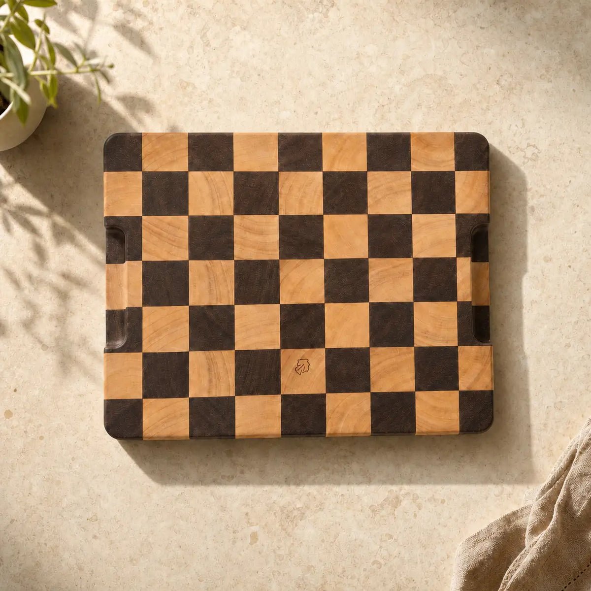 ALORIVO end-grain Sapele wooden cutting board with a luxury checkerboard pattern, combining dark Sapele wood and light wood blocks. The board features the ALORIVO logo etched near the bottom center, displayed on a beige stone kitchen counter next to a linen cloth.