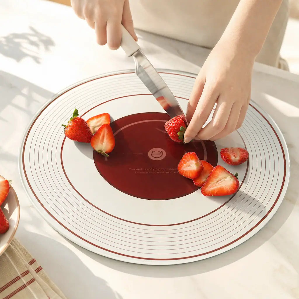 Close-up of a chef slicing fresh red strawberries with a white knife on the ALORIVO circular tempered glass cutting board featuring a vinyl record graphic with concentric grooves, bathed in bright California sunlight on a marble countertop.